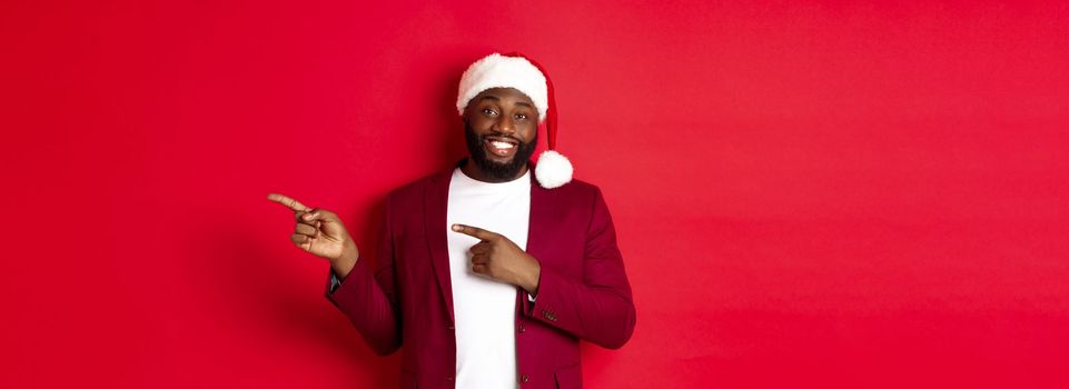 Christmas, party and holidays concept. Smiling Black man with beard and santa hat, pointing fingers right at showing logo, standing over red background.
