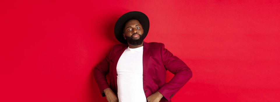 Funny african american man trying to act serious, holding hands on waist and staring at camera scary, standing over red background.