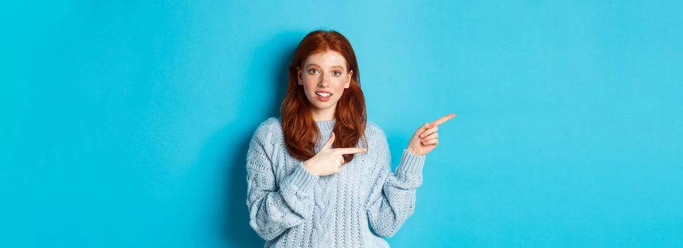 Image of pretty redhead girl in sweater, pointing fingers left at logo, smiling curious, standing over blue background.