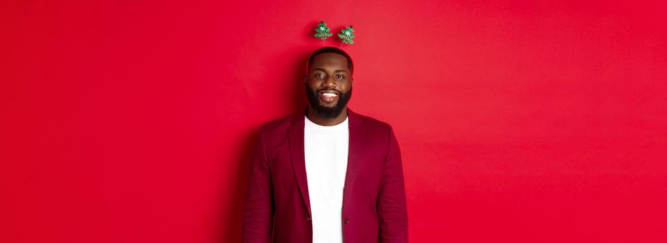 Merry Christmas. Happy african american man celebarting New Year, wearing funny party headband and smiling, standing over red background.