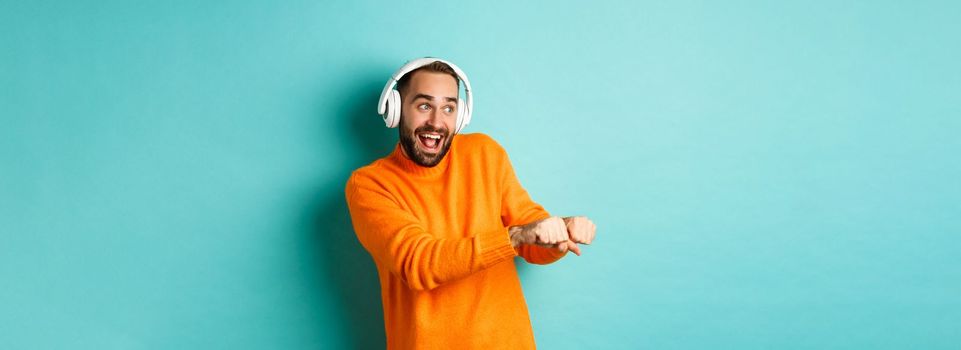 Happy man listening music in headphones and dancing funny, standing over turquoise background. Copy space