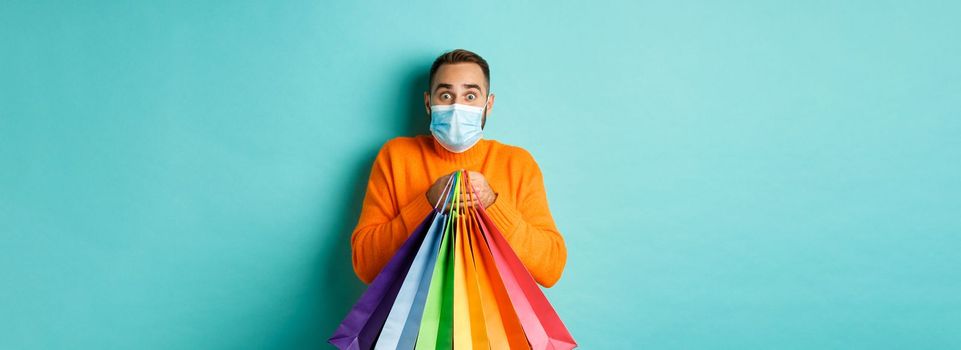 Covid-19, pandemic and lifestyle concept. Excited man in face mask showing shopping bags and rejoicing from discounts, standing over turquoise background.