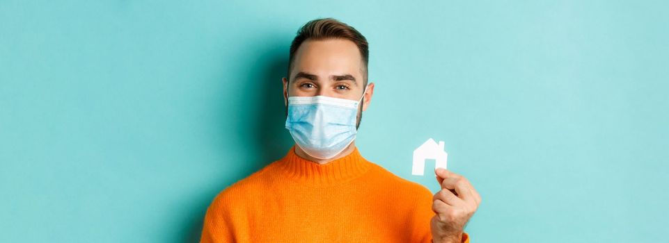 Real estate and coronavirus pandemic concept. Close-up of adult man in medical mask holding small paper house maket and smiling, searching for apartment, light blue background.