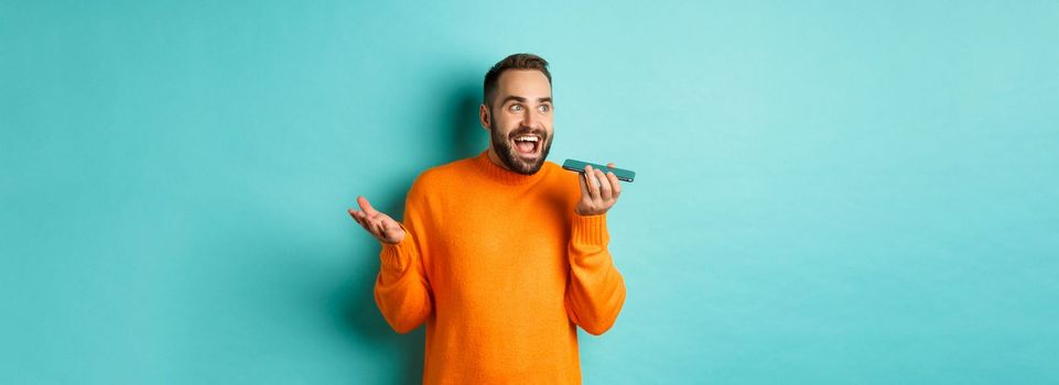 Happy man talking on speakerphone, gesturing and recording voice message on mobile phone, standing in orange sweater over light blue background.