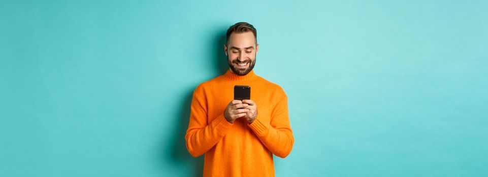Handsome man smiling and texting message on mobile phone, communicating online, standing over light blue background.