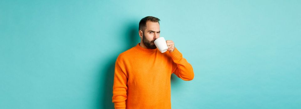 Image of guy drinking coffee and looking suspicious at camera, stare at something strange, standing in orange sweater over turquoise background.