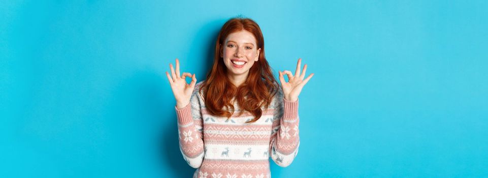 Cute redhead girl in sweater showing okay signs, smiling pleased, approve and like, praise excellent choice, standing over blue background.