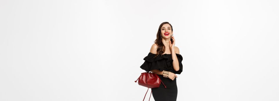 Beauty and fashion concept. Full length of elegant young woman in black cocktail dress, holding purse and wearing makeup, laughing at camera, standing over white background.