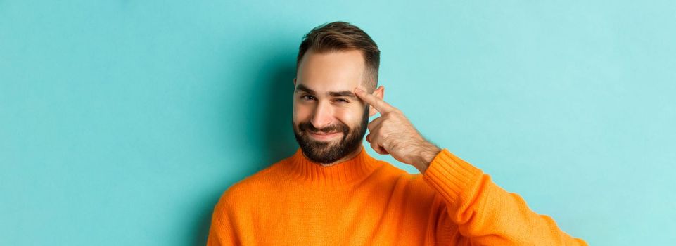 Image of young man in orange sweater, smiling and pointing at head, having an idea, praising good thought, standing over light blue background.