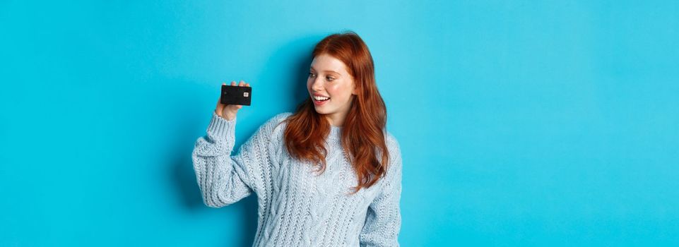 Cute redhead girl in sweater showing credit card, smiling at camera, standing over blue background.