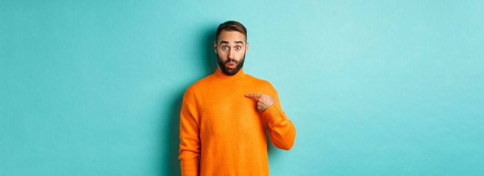 Man pointing at himself with surprise face, being chosen, standing confused against light blue background.