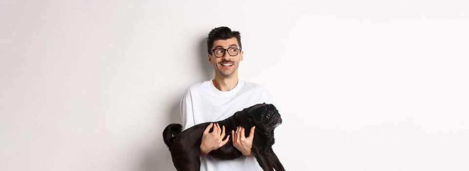 Excited hipster guy with his dog looking left at logo. Handsome pet owner carry black pug and staring at promo banner, standing over white background.