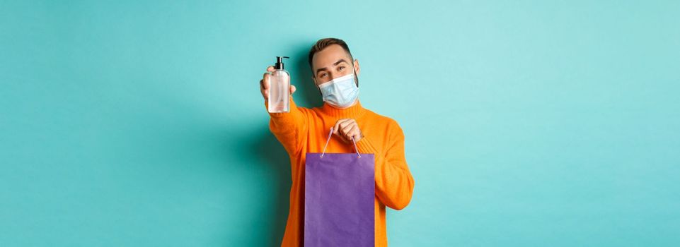 Coronavirus, pandemic and lifestyle concept. Man in face mask showing shopping bag and hand sanitizer, standing over turquoise background.