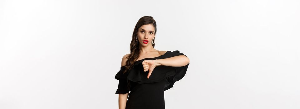 Fashion and beauty. Disappointed sassy woman in black dress, showing thumbs down, dislike something bad, judging over white background.