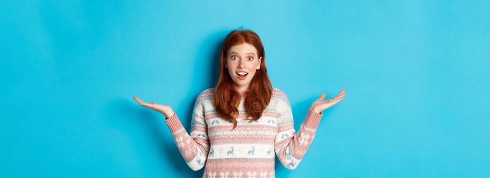 Image of excited redhead girl react to good news, looking surprised, spread hands sideways and smiling, standing in winter sweater against blue background.