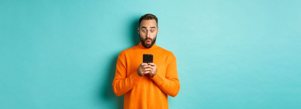 Image of man reading message with surprised face, standing amazed over light blue background.