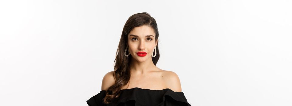 Fashion and beauty concept. Close-up of elegant brunette woman with earrings, wearing black dress and red lipstick, looking sensual at camera, standing over white background.