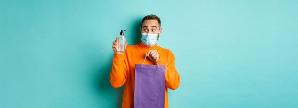 Coronavirus, pandemic and lifestyle concept. Man in face mask showing shopping bag and hand sanitizer, standing over turquoise background.