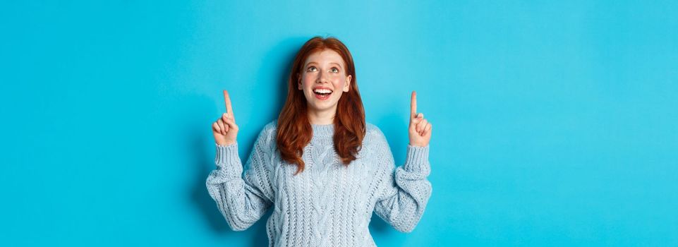 Winter holidays and people concept. Beautiful teenage girl with red hair, wearing sweater, pointing fingers up at logo and smiling, showing advertisement, blue background.