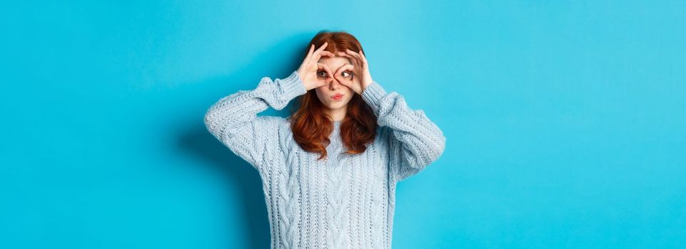 Funny redhead female model in sweater, staring at camera through fingers glasses, express interest and amazement, standing over blue background.