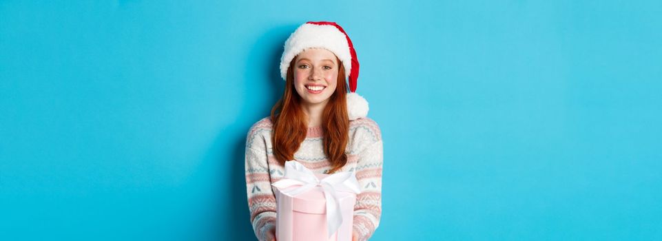 Winter and celebration concept. Beautiful redhead girl in santa hat wishing merry christmas, giving gift and smiling, standing over blue background.