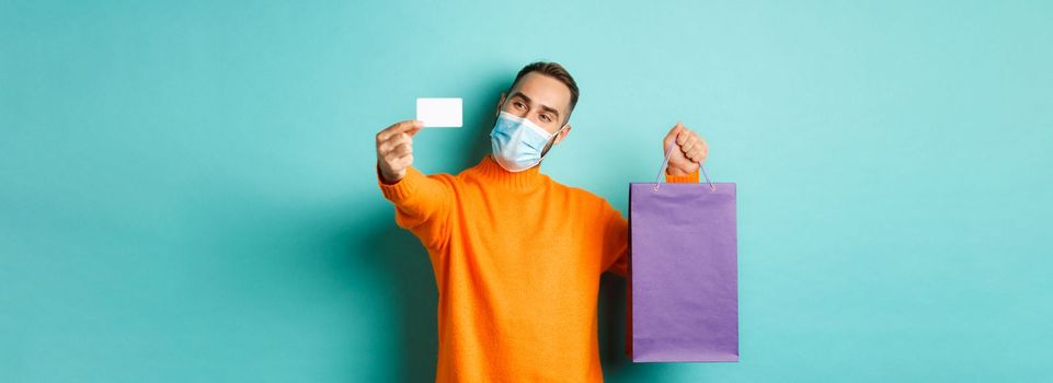 Covid-19, pandemic and lifestyle concept. Happy male customer in face mask showing credit card and purple shopping bag, standing over light blue background.