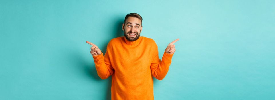 Confused handsome man shrugging indecisive, pointing fingers sideways, cant choose between two variants, standing against light blue background.