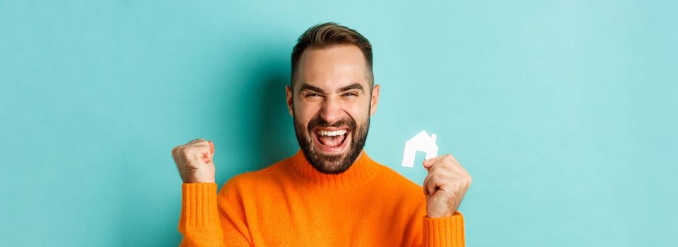 Real estate. Cheerful man buying apartment, rejoicing and saying yes, showing small paper house, standing over light blue background.