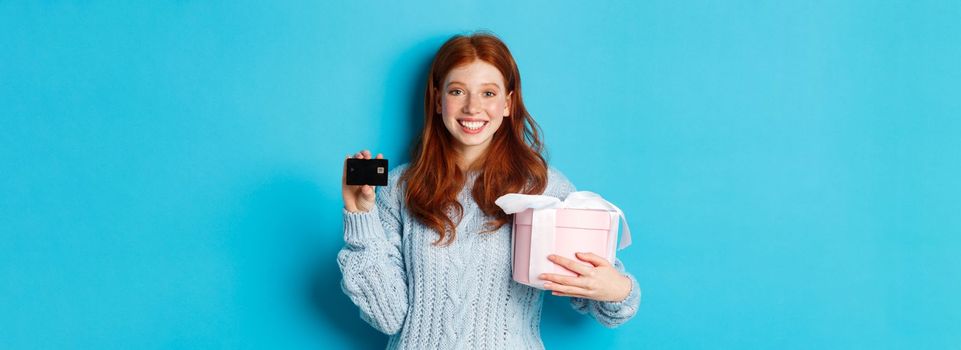 Happy redhead girl buying gifts with credit card, holding box with present and smiling, standing over blue background.