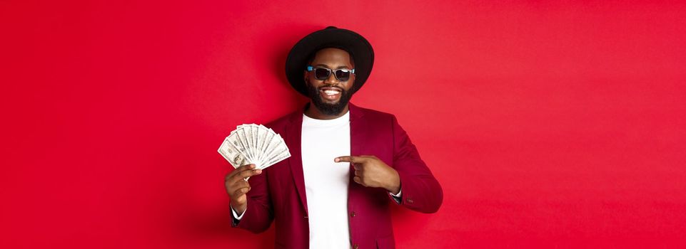 Handsome and stylish african american male model showing money and smiling, wearing sunglasses and fancy hat, standing over red background.