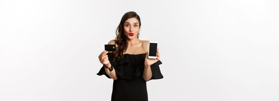 Fashion and online shopping concept. Happy young woman in black dress, showing credit card and mobile screen, standing over white background.