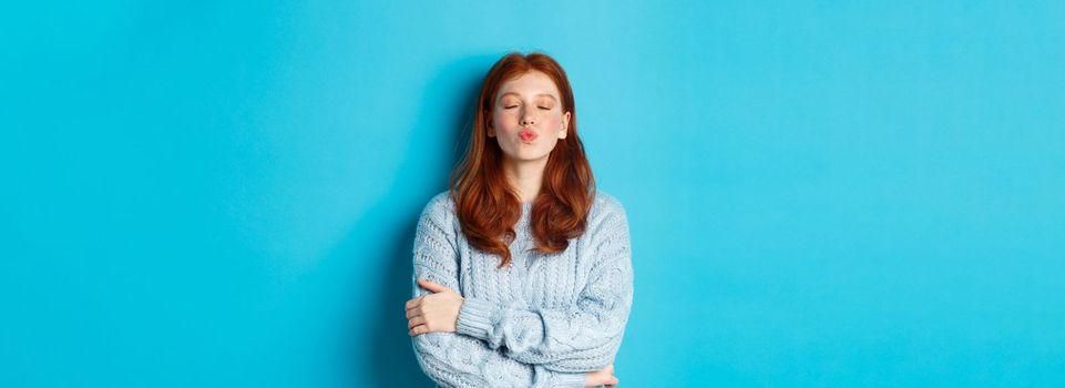 Cute redhead teen girl waiting for kiss, pucker lips and close eyes, standing in sweater against blue background.