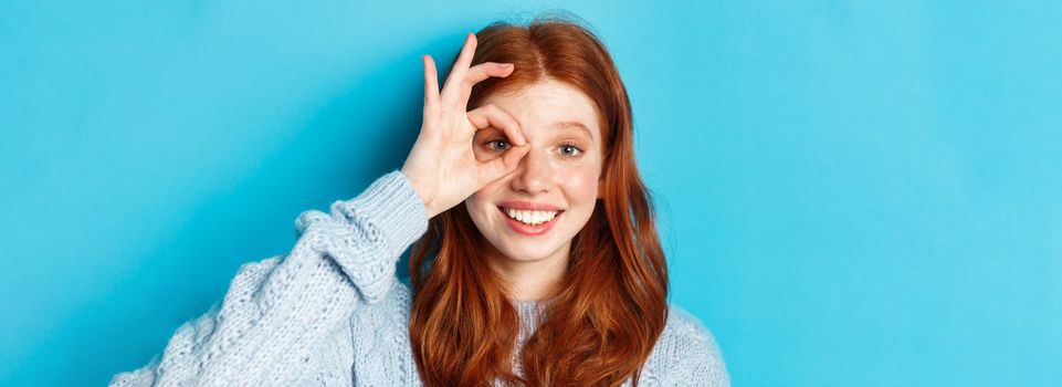 Headshot of cute teenage redhead girl showing okay sign on eye and smiling, standing happy against blue background.