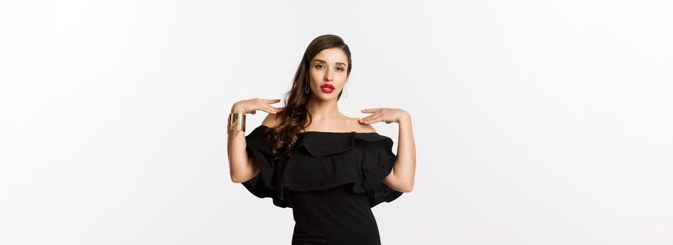 Confident and stylish woman in black elegant dress, looking sassy at camera, standing over white background.