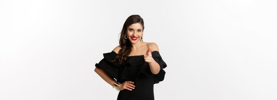 Fashion and beauty. Sassy pretty woman in black dress and makeup, pointing finger at camera to congratulate or praise, standing over white background.