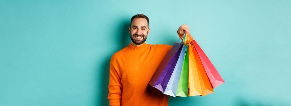 Happy handsome man holding shopping bags and smiling, buying presents, standing over turquoise background.