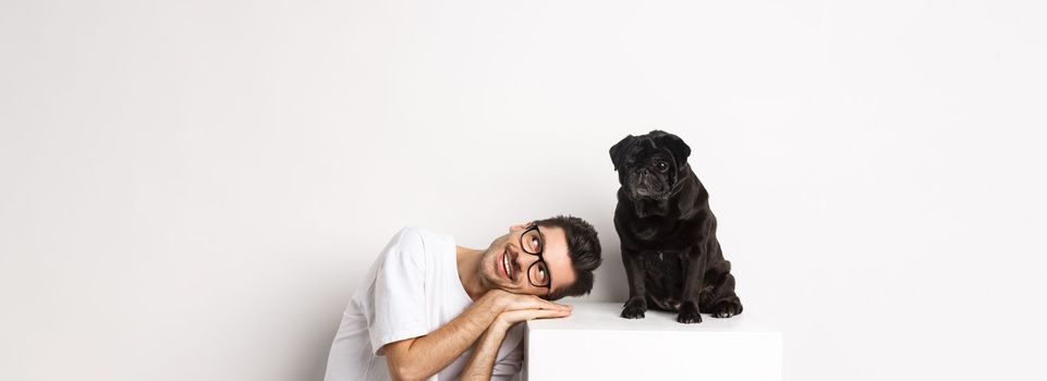 Handsome young man lay head near cute black pug, smiling and looking up at copy space, white background.