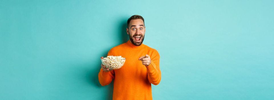 Handsome young man holding bowl of popcorn, smiling amazed and pointing at camera, standing over blue background.