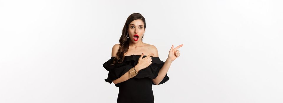 Fashion and beauty. Surprised woman in black glamour dress pointing fingers left, showing advertisement and staring amazed, white background.
