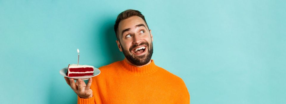 Close-up of happy adult man celebrating birthday, holding bday cake with candle and making wish, standing against turquoise background.