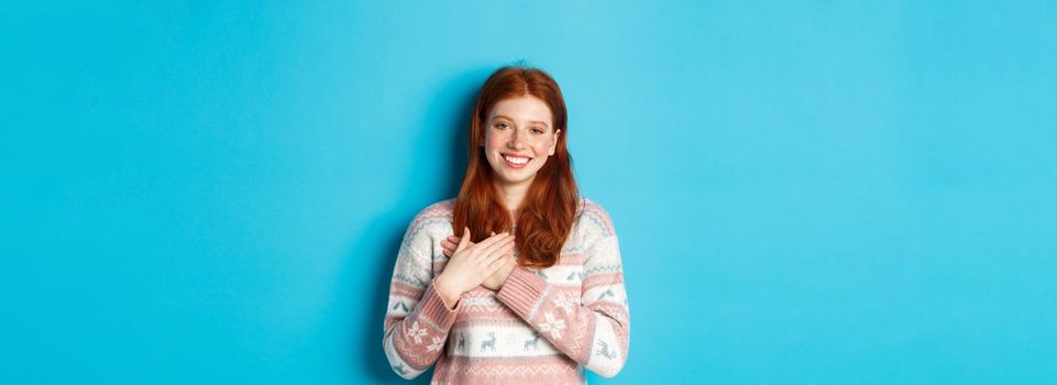 Image of beautiful redhead female model holding hands on heart and smiling, saying thank you, being grateful, standing over blue background.