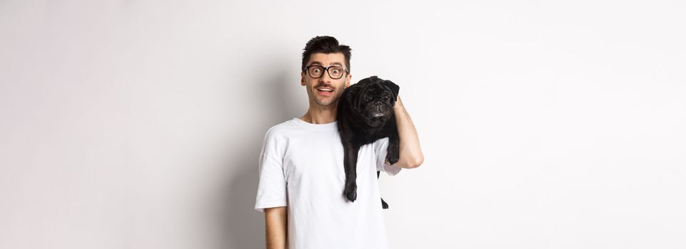 Amazed young man in glasses holding black pug on shoulder and staring at camera impressed. Dog owner posing with cute puppy near white background.