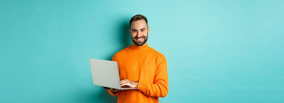 Handsome adult man freelancer working with laptop, smiling at camera, typing on computer keyboard, standing over light blue background.