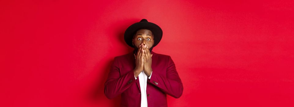 Fashion and party concept. Shocked Black man react on christmas advertisement, gasping and staring at camera with disbelief, standing over red background.