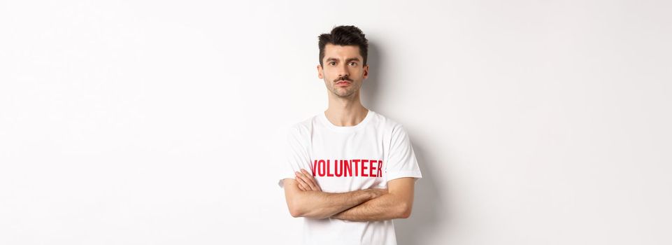 Serious young male volunteer in white t-shirt, holding arms crossed on chest, looking at camera, ready to help.