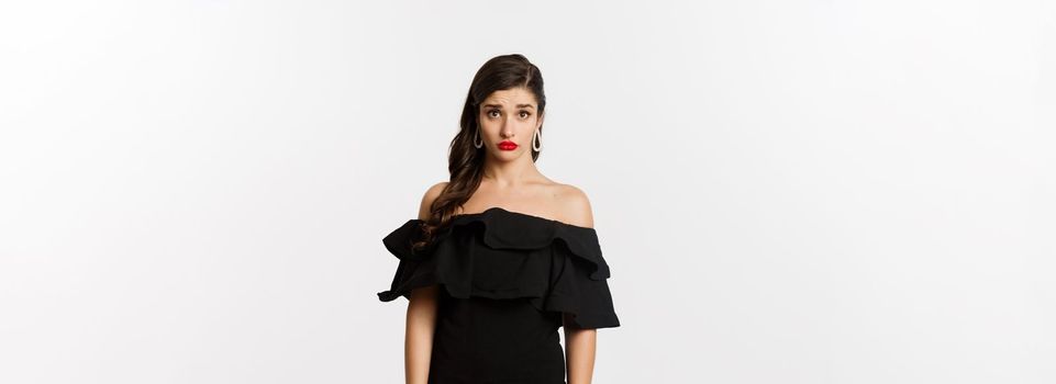 Fashion and beauty. Cute and timid young woman in black dress looking confused and sad at camera, cant understand, standing gloomy against white background.