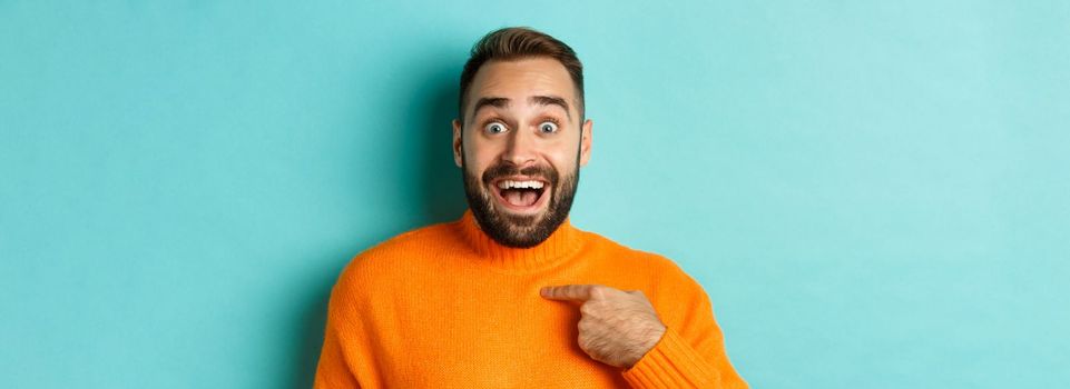 Close-up of happy young man pointing at himself with surprise and excitement, standing over light blue background.