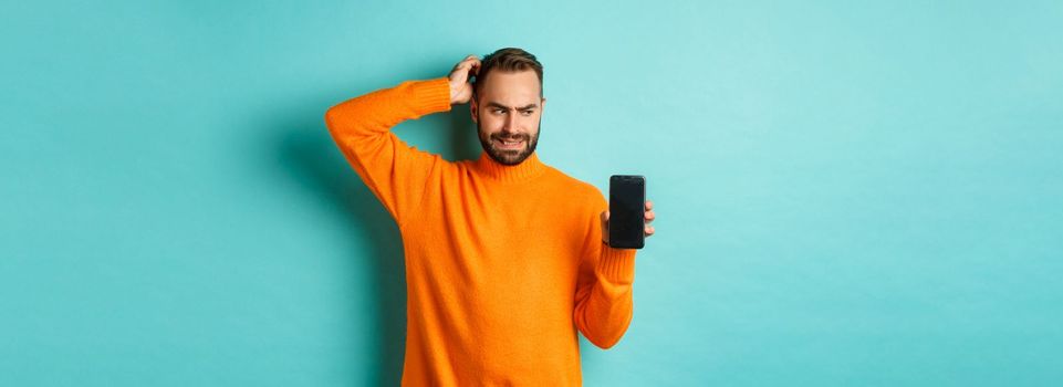 Confused guy scartching head and showing smartphone screen, looking puzzled, standing over turquoise background. Copy space