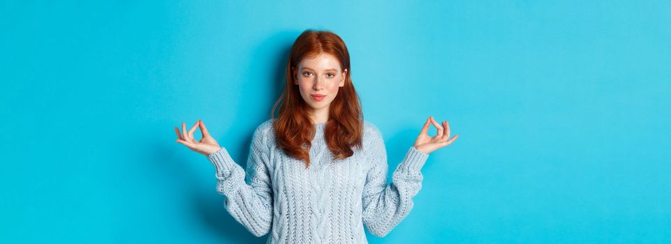 Smiling confident girl with red hair staying patient, holding hands in zen, meditation pose and staring at camera, practice yoga, standing calm against blue background.