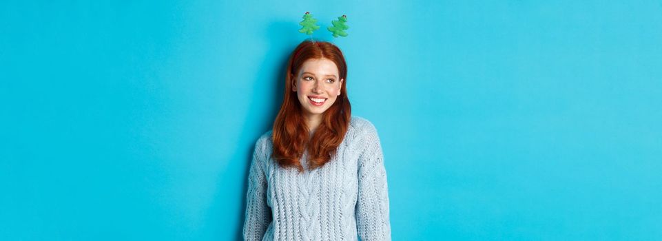 Winter holidays and Christmas sales concept. Cute redhead girl in funny New Year headband smiling, looking left at logo, standing over blue background.